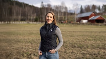 Anna Östlund står på ett fält med ladugårdsbyggnad i bakgrunden