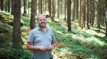 Leende man i ljusgrå pikétröja står i en solbelyst skog med mossa och höga träd i bakgrunden.