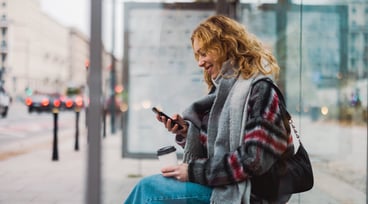 Kvinna väntar på bussen och tittar ner i sin mobiltelefon.