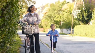 Mamma och son på promenad i villakvarter. Mamman leder en cykel och sonen åker på en sparkcykel.
