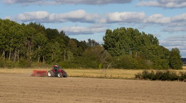 Traktor kör på åker. Blå himmel och skog i bakgrunden.