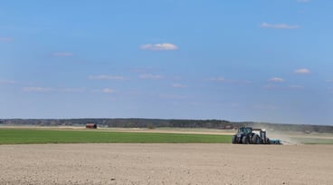 Vy över åker och blå himmel, traktor och såmaskin på avstånd
