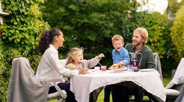 Familj fikar i trädgården tillsammans. Gröna buskar och träd i bakgrunden.