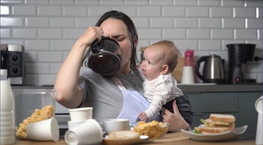 Mamma dricker kaffe direkt ur kannan vid köksbordet med sin bebis i famnen
