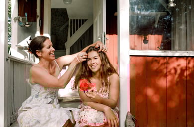 Mor och dotter sitter utomhus. Mamma fixar med dotterns hår.