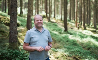 Leende man i ljusgrå pikétröja står i en solbelyst skog med mossa och höga träd i bakgrunden.