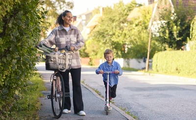 Mamma och son på promenad i villakvarter. Mamman leder en cykel och sonen åker på en sparkcykel.
