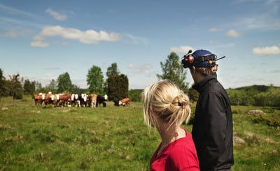 Två lantbrukare, en kvinna och en man, står med ryggen mot kameran och tittar ut över en kohage