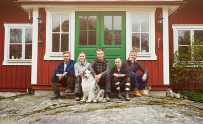 Familj sitter utanför entrén  till ett rött hus med gröna dubbeldörrar.