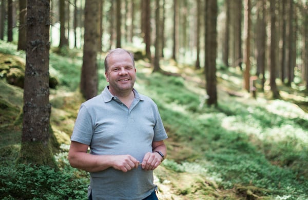 Leende man i ljusgrå pikétröja står i en solbelyst skog med mossa och höga träd i bakgrunden.