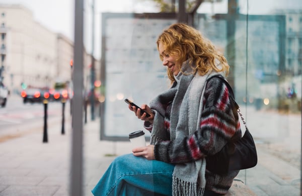 Kvinna väntar på bussen och tittar ner i sin mobiltelefon.