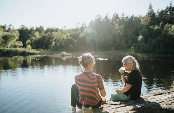 Två äldre kvinnor sitter på en klippa vid vattnet och fikar.
