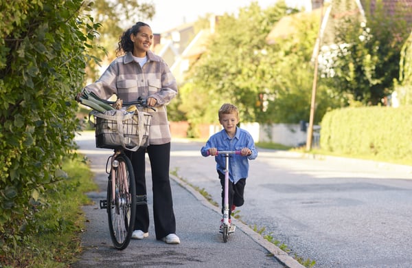 Mamma och son på promenad i villakvarter. Mamman leder en cykel och sonen åker på en sparkcykel.