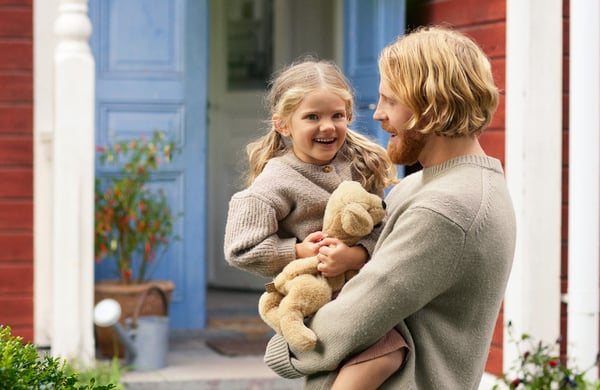 En pappa håller sin dotter i famnen. Rött hus med blå dörr skymtar i bakgrunden. Gröna buskar i förgrunden.